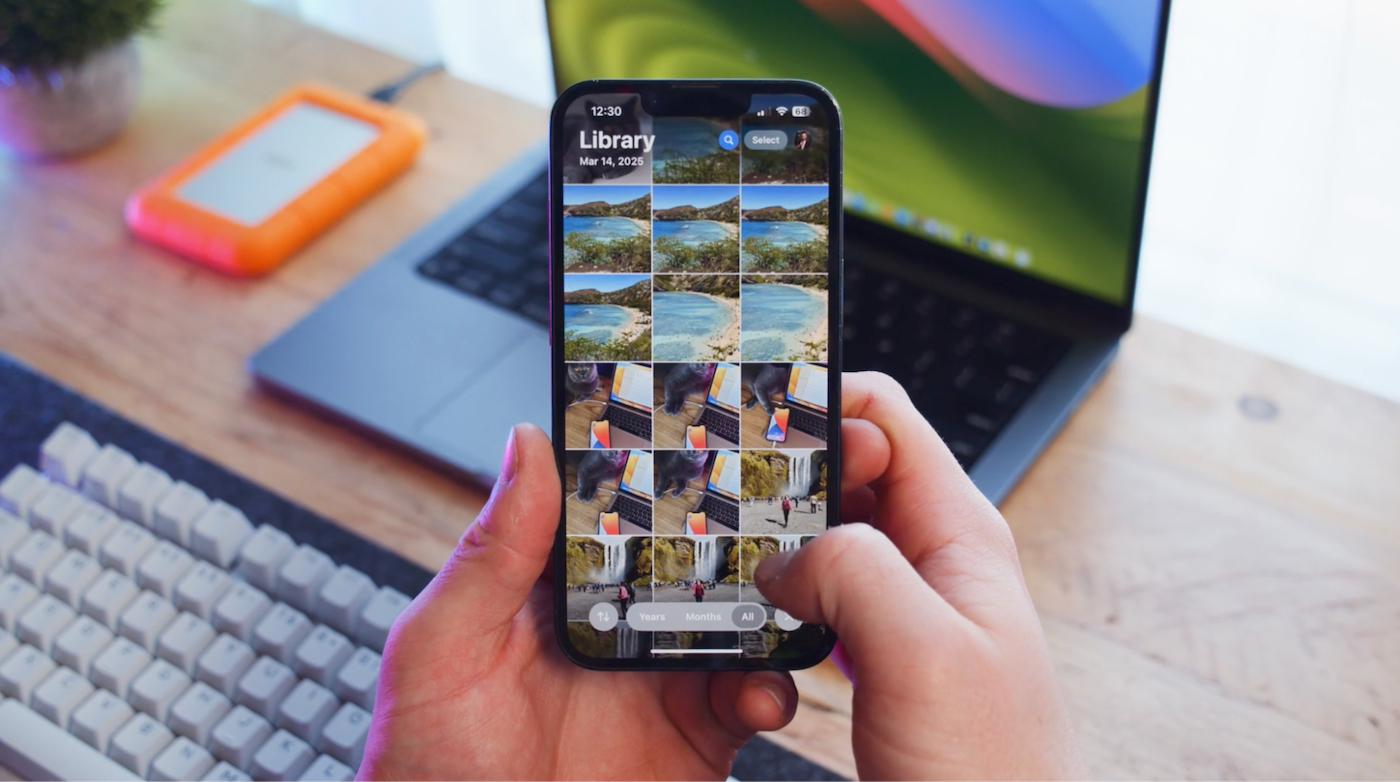 Person organizing photo library on iPhone to free up storage space, with laptop and external drive in the background