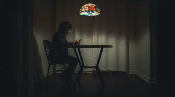 Man sitting alone in a dark room looking at his phone under a lamp